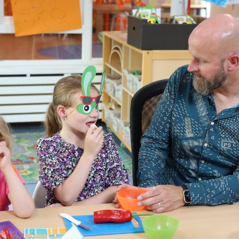 Wethouder TRick Vermin van de gemeente 's-Hertogenbosch aan tafel met een meisje dat met een konijnenmasker op groenten proeft 