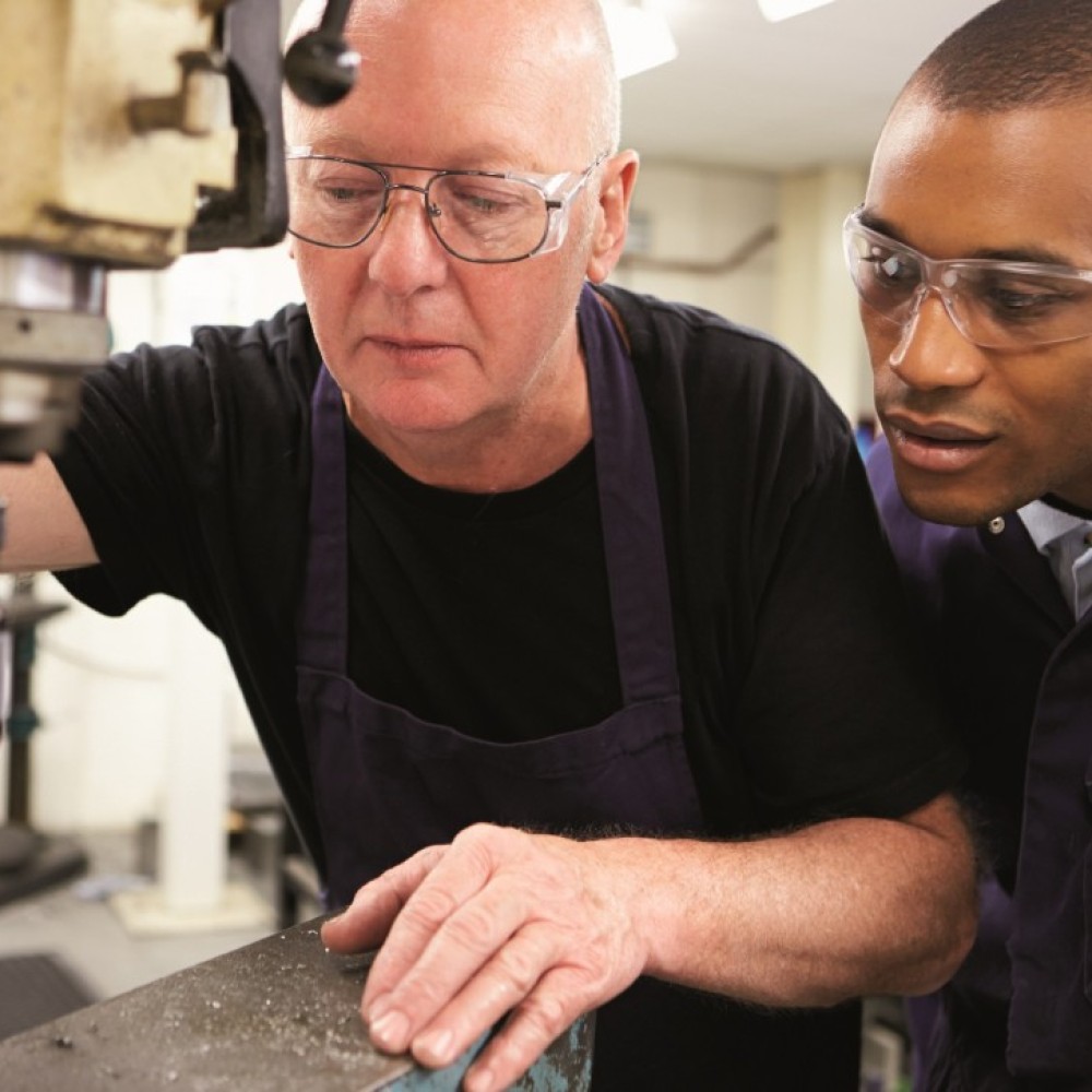 Een oudere fabrieksmedewerker helpt een jongere collega bij een metaalmachine