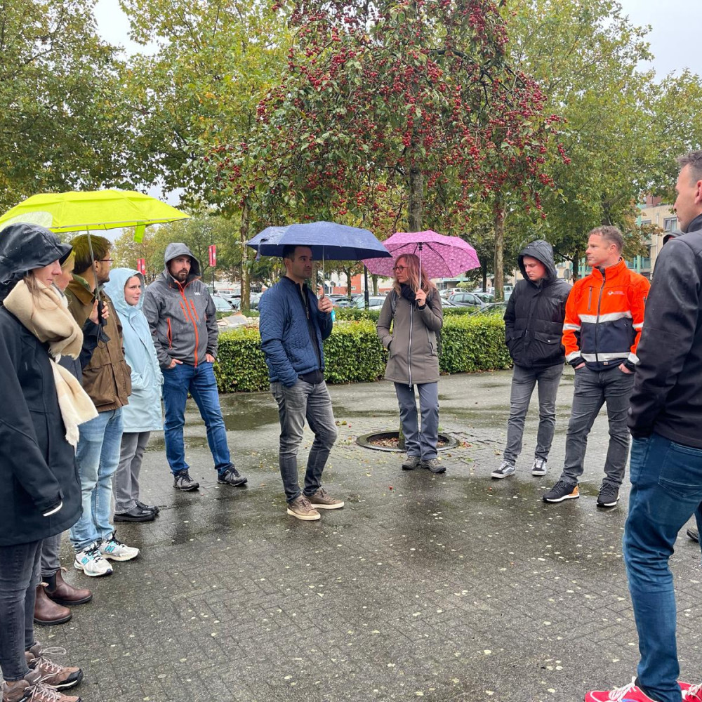 Groepje van 10 mensen staan buiten bij een parkeerplaats op een rij, sommigen met een paraplu in de hand