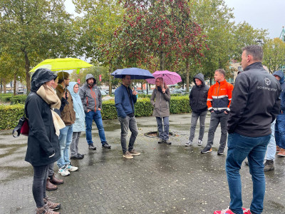 Groepje van 10 mensen staan buiten bij een parkeerplaats op een rij, sommigen met een paraplu in de hand