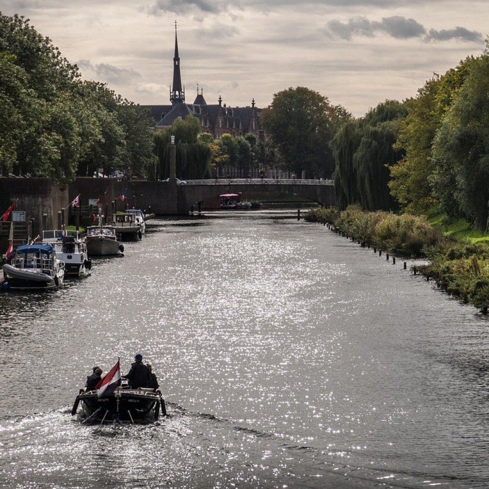 Klein bootje met Nederlandse vlag vaart over een rivier richting een brug in Den Bosch, omringd door bomen en gebouwen met een kerktoren op de achtergrond.
