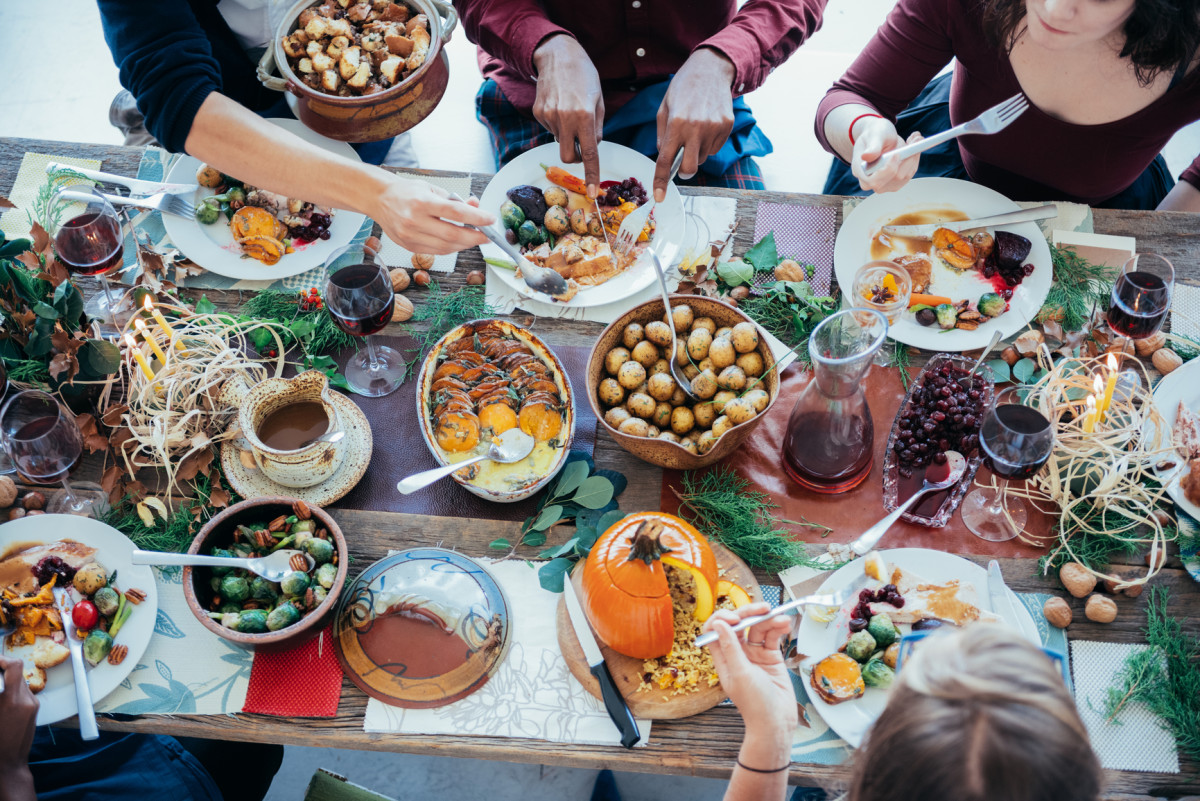 Foto van een groepje mensen aan een eettafel van bovenaf genomen, met gerechten zoals aardappelen, pompoen en groenten; er staan een karaf glazen rode wijn op tafel en iedereen is aan het eten of opscheppen.