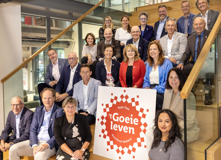Groepsfoto van de eerste ambassadeurs van de Regio Deal ’t Goeie Leven in Noordoost-Brabant. Zij staan en zitten op een trap in een binnenruimte, met een groot bord met het logo van ’t Goeie Leven op de voorgrond