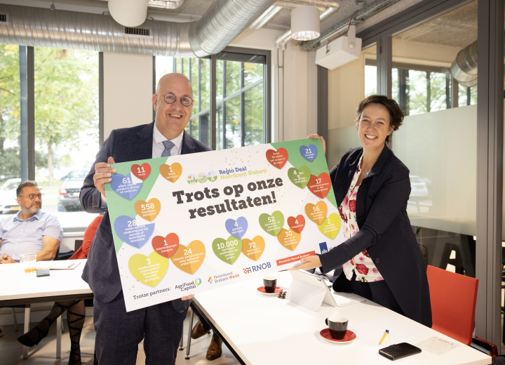 Foto waarop de voorzitter van de vorige regio deal de opbrengst van de deal uitreikt aan de programmamanager Regio Deals van BZK op een groot bord met hartjes en  een opsomming van resultaten.