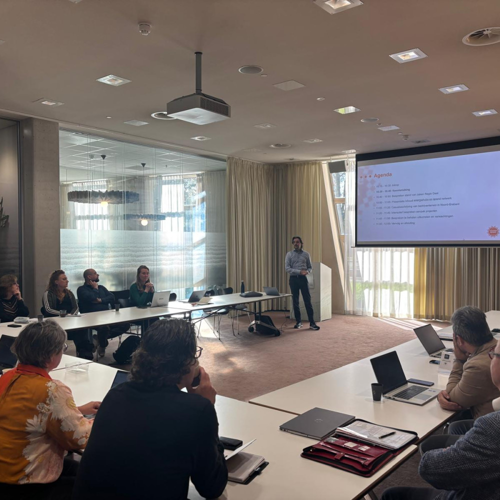 Projectleider presenteert voor een scherm aan mensen in een zaal die aan een tafel in U-vorm zitten met laptops en notitieblokken