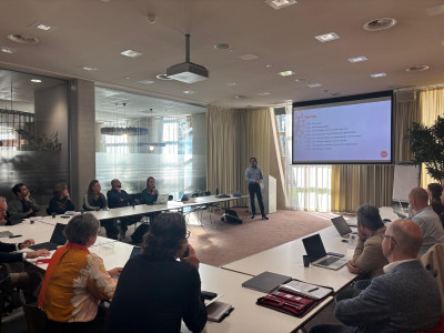 Projectleider presenteert voor een scherm aan mensen in een zaal die aan een tafel in U-vorm zitten met laptops en notitieblokken