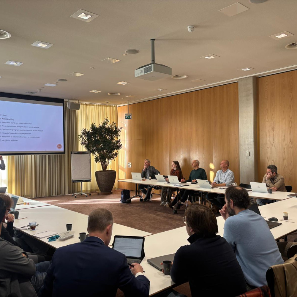 Deelnemers aan de start bijeenkomst zitten aan een tafel in U-vorm met laptops en schrijfmateriaal