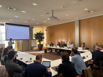 Deelnemers aan de start bijeenkomst zitten aan een tafel in U-vorm met laptops en schrijfmateriaal
