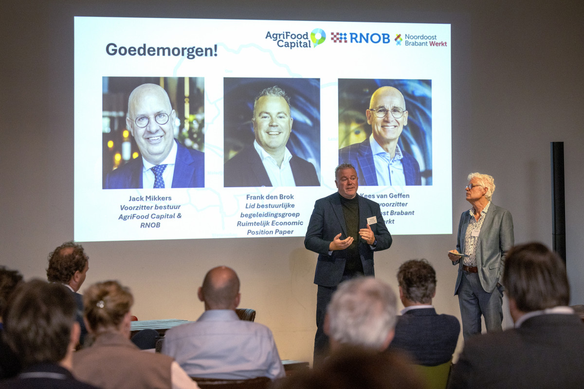 Wethouder Frank den Brok samen met de dagvoorzitter voor een scherm met namen van sprekers en de logo’s van hun organisaties.