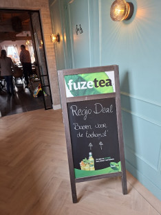 Een groot krijtbord op de grond met een welkomsttekst en pijlen als wegwijzers voor de deelnemers aan de bijeenkomst. Op de bovenkant reclame voor het drankje Fuzetea.