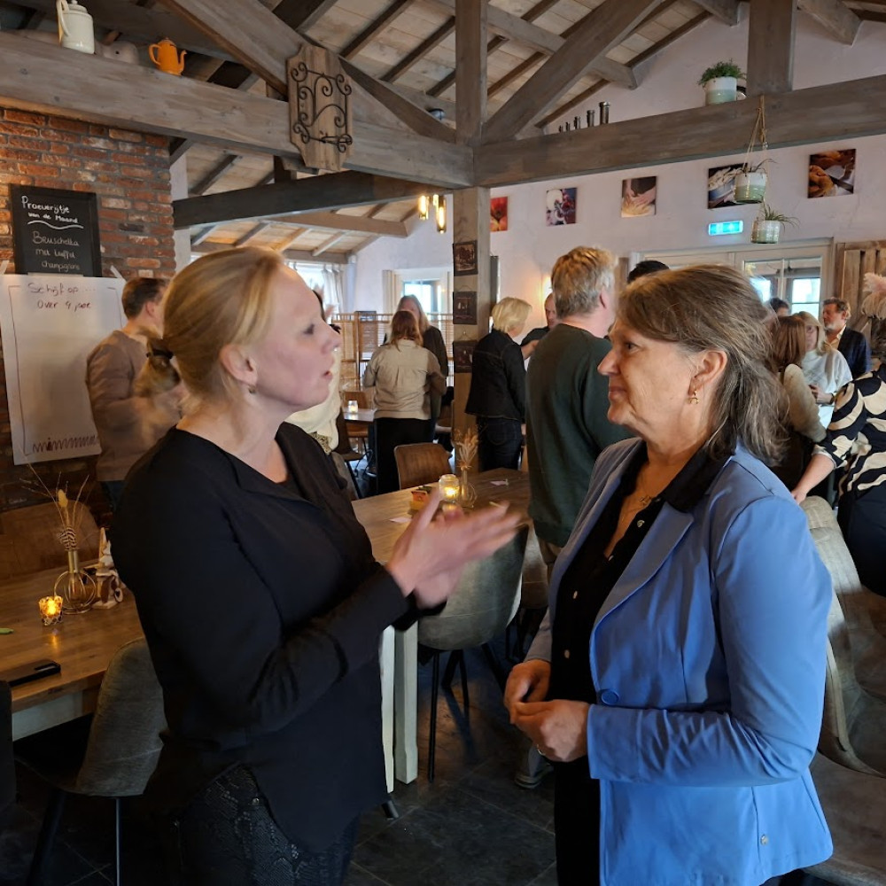 Op de voorgrond twee deelnemers van de bijeenkomst met elkaar in gesprek voor een houten tafel. Op de achtergrond praten andere aanwezigen met elkaar. 