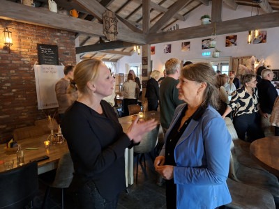 Op de voorgrond twee deelnemers van de bijeenkomst met elkaar in gesprek voor een houten tafel. Op de achtergrond praten andere aanwezigen met elkaar. 