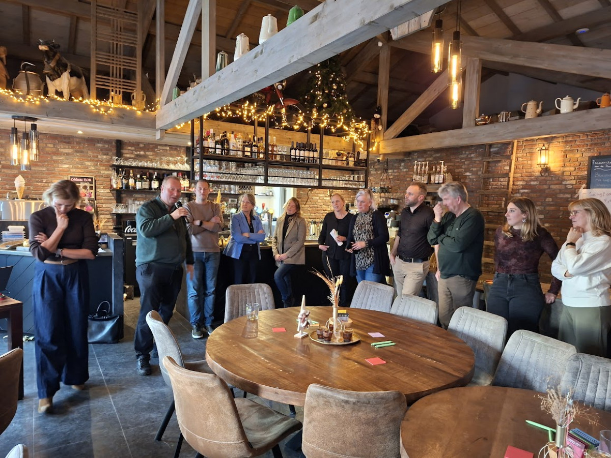 Deelnemers bijeenkomst in een sfeervol restaurant met houten balken, warme verlichting en kerstdecoratie. Ze staan in een halve cirkel bij de bar, terwijl een spreker iets presenteert. Op de voorgrond een ronde houten tafel met decoratieve vaas en kaartjes.