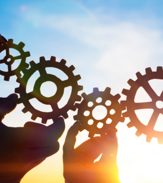 Drie tandwielen die in elkaar grijpen, vastgehouden door handen, tegen een achtergrond van een heldere lucht met zonlicht. Dit beeld symboliseert samenwerking en efficiëntie.