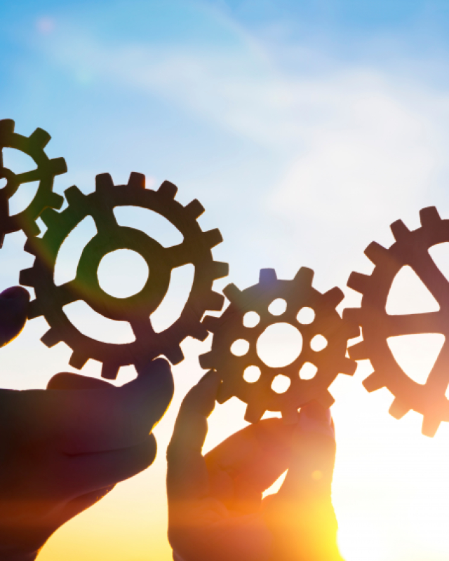 Drie tandwielen die in elkaar grijpen, vastgehouden door handen, tegen een achtergrond van een heldere lucht met zonlicht. Dit beeld symboliseert samenwerking en efficiëntie.