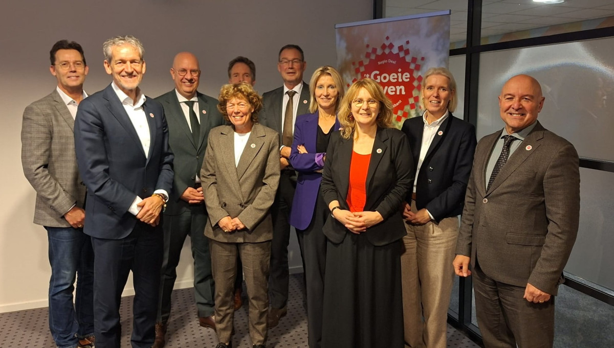 De leden van de Algemene Vergadering RNOB staan naast elkaar op ene rij in een vergaderruimte  vóór een rollup banner met het logo van de Regio Deal.