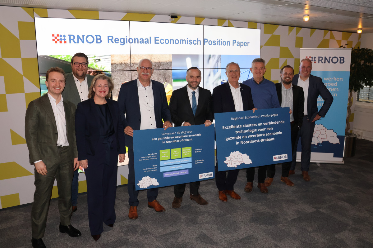 Groepsfoto van wethouders uit de regio Noordoost-Brabant. Ze houden een bord met een tekst en afbeelding vast en staan vóór een groot scherm met teksten en afbeeldingen en een rollup banner van RNOB