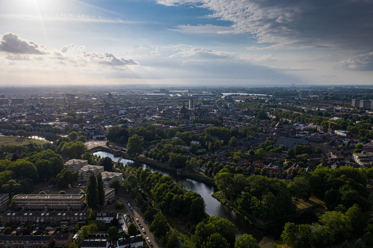 Foto van de stad Den Bosch van bovenaf met zichtbare bomen, wegen, huizen, de Sint Jan en water onder een blauwe lucht met zon en met grijze wolken