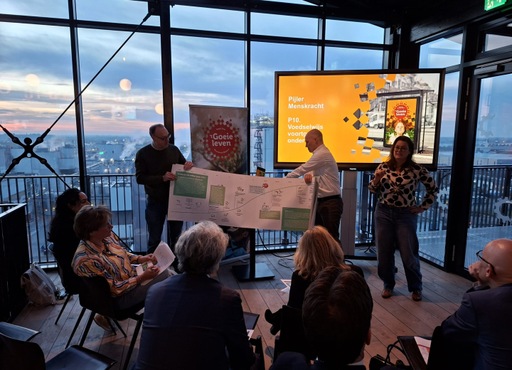 Presentatie van projectplannen tijdens een bijeenkomst van de Stuurgroep Regio Deal bij The Chocolate Factory met uitzicht over de stad. De sprekers lichten een groot schema toe die ze vasthouden.