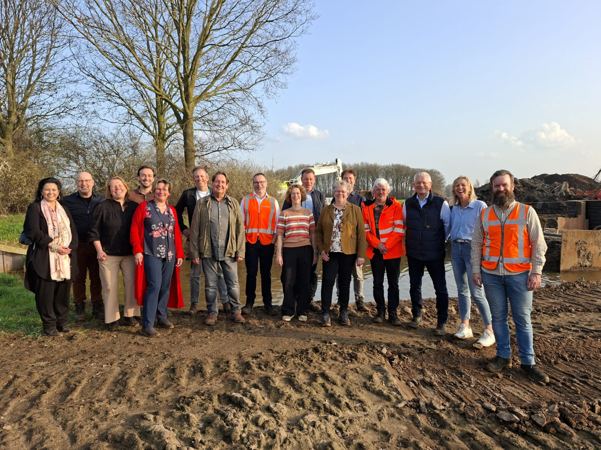 Groepsfoto van alle deelnemers van de startbijeenkomst in het zand vóór het werkterrein met een kraan op de achtergrond