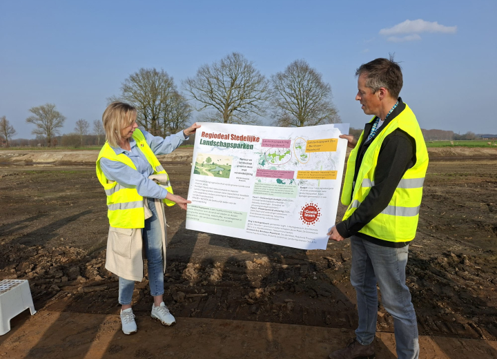 Twee deelnemers in veiligheidshesjes presenteren een groot informatiepaneel over gebiedsontwikkeling op de projectlocatie in het buitengebied
