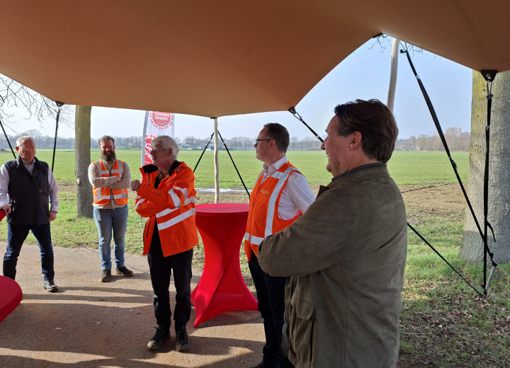De deelnemers aan de bijeenkomst met veiligheidshesjes onder een tentdoek in een landelijk gebied.