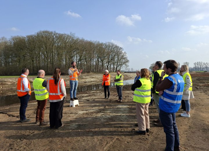 Groep deelnemers met veiligheidshesjes tijdens een uitlegmoment op de projectlocatie voor water- en gebiedsontwikkeling in een landelijk gebied.
