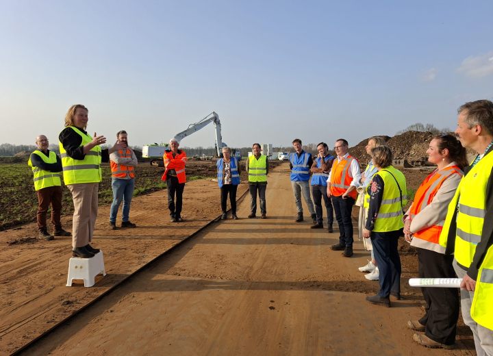 Groep deelnemers met veiligheidshesjes tijdens een uitlegmoment op de projectlocatie voor water- en gebiedsontwikkeling in een landelijk gebied met een kraan op de achtergrond