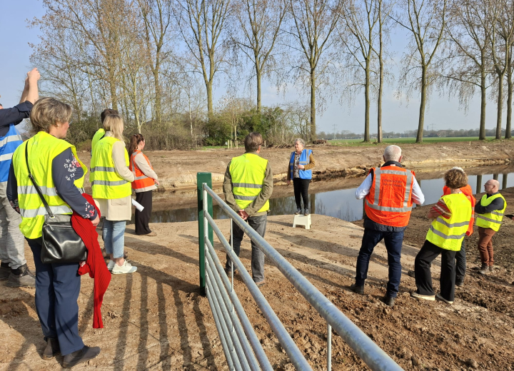 Groep deelnemers met veiligheidshesjes tijdens een uitlegmoment op de projectlocatie voor water- en gebiedsontwikkeling in een landelijk gebied