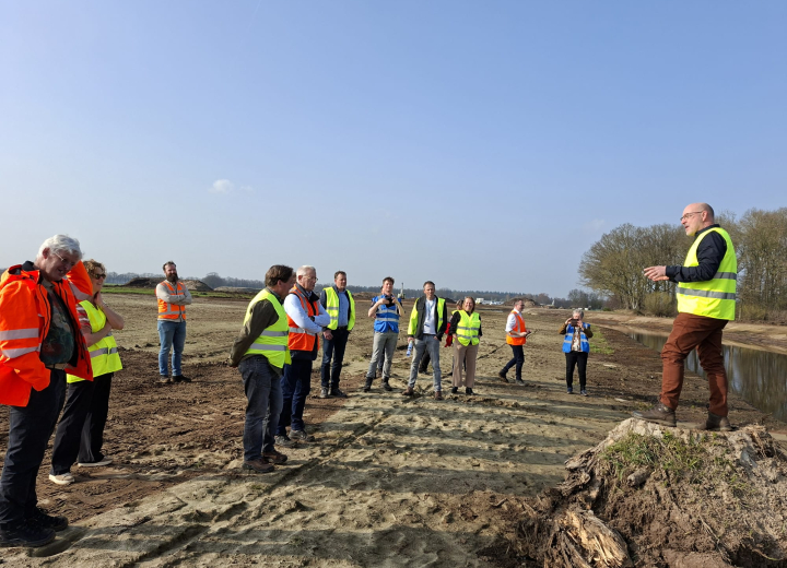 Groep deelnemers met veiligheidshesjes tijdens een uitlegmoment op de projectlocatie voor water- en gebiedsontwikkeling in een landelijk gebied