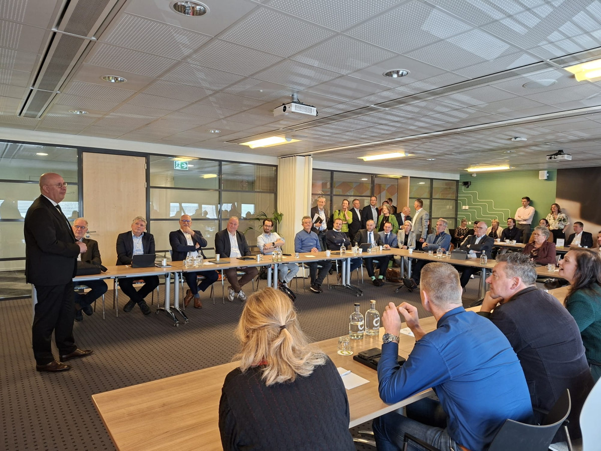 Wethouders aan tafel in een grote zaal aan een tafel in U-vorm met Jack Mikkers voor in de zaal die iedereen toespreekt