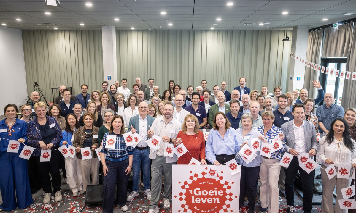 Groepsfoto van alle aanwezigen van de feestelijke startbijeenkomst van Regio Deal 't Goeie leven.  Ze staan achter een groot bord met het Regio Deal logo erop en de mensen op de eerste rij houden vlaggetjes met het logo vast.