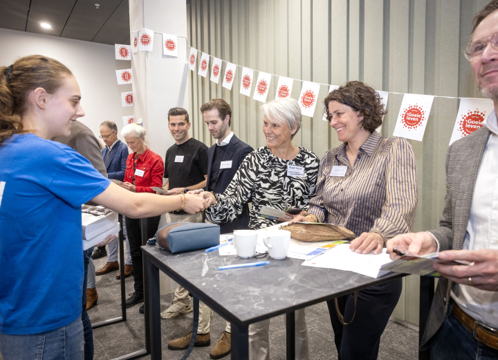 Een student reikt aan deelnemers aan het startevent een speldje uit met het Regio Deal logo.