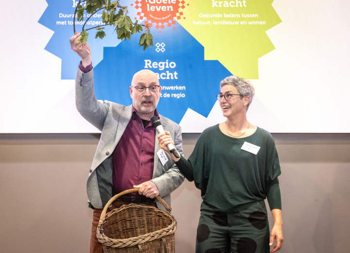 Twee programmamanagers van de Regio Deal lichten de inhoud van hun programma's toe, met een mand groenten op het podium met op de achtergrond een illustratie