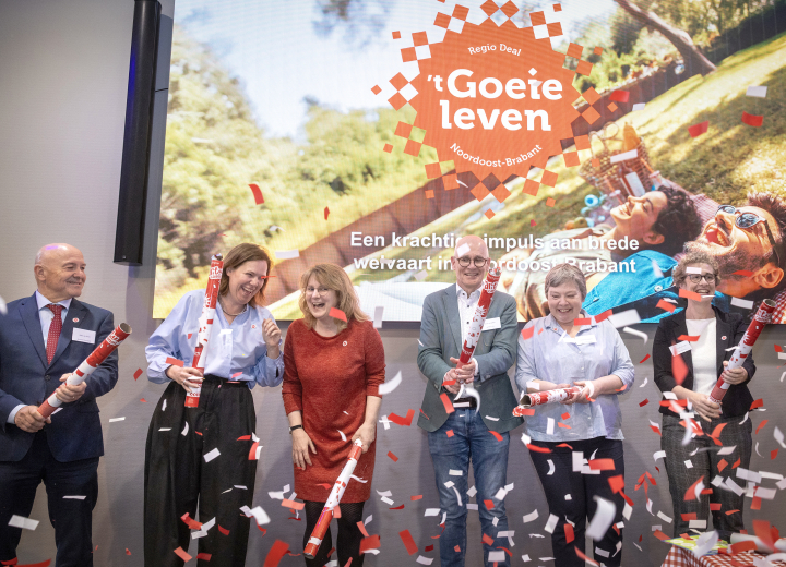 Feestelijk moment tijdens Regio Deal startbijeenkomst bestuurders met confetti op het podium. Op de achtergrond een foto met het Regio Deal logo.