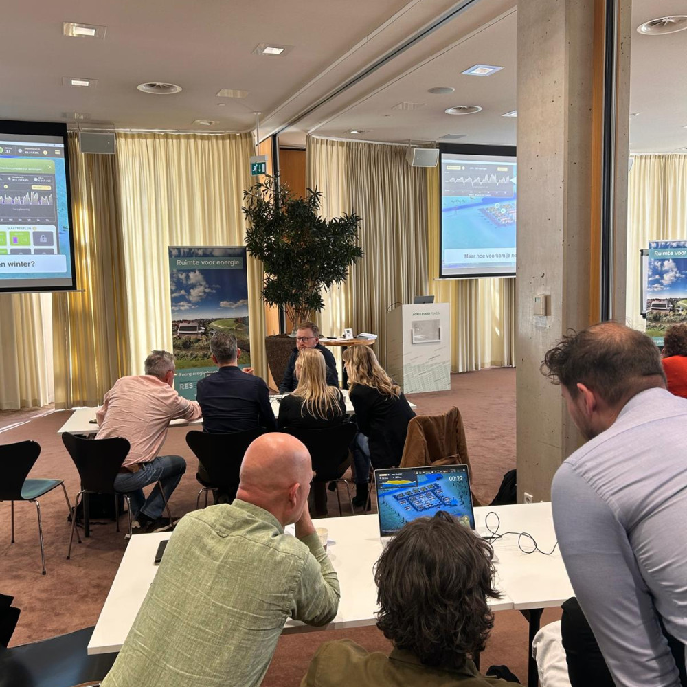 Deelnemers aan de masterclass werken in een grote vergaderzaal in groepjes aan tafels met laptops aan opdrachten. Op de achtergrond zijn presententatieschermen zichtbaar.