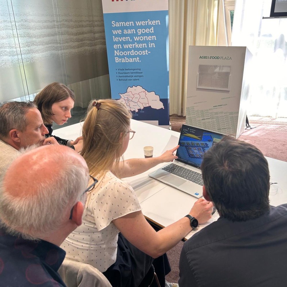 Een groepjke deelnemers aan de masterclass werkt in een grote vergaderzaal aan een tafel met een laptop aan opdrachten. Op de achtergrond staat een rollup banner van RNOB.