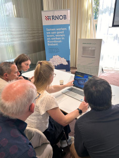 Een groepjke deelnemers aan de masterclass werkt in een grote vergaderzaal aan een tafel met een laptop aan opdrachten. Op de achtergrond staat een rollup banner van RNOB.