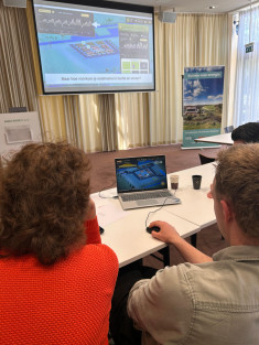Deelnemers spelen een serious game op een laptop tijdens de workshop met op de achtergrond een presentatie.