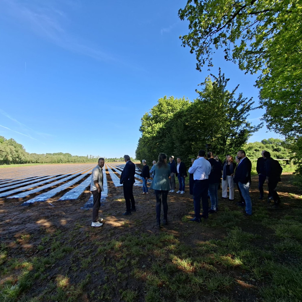 Groep wethouders staan op een landbouwveld met afgedekte rijen gewassen met bomen op de achtergrond.