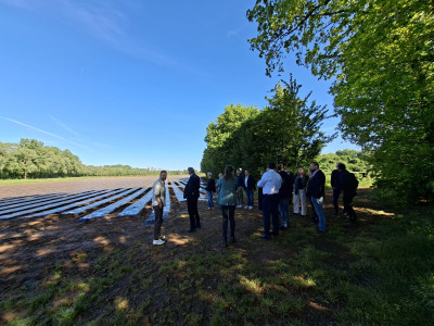 Groep wethouders staan op een landbouwveld met afgedekte rijen gewassen met bomen op de achtergrond.