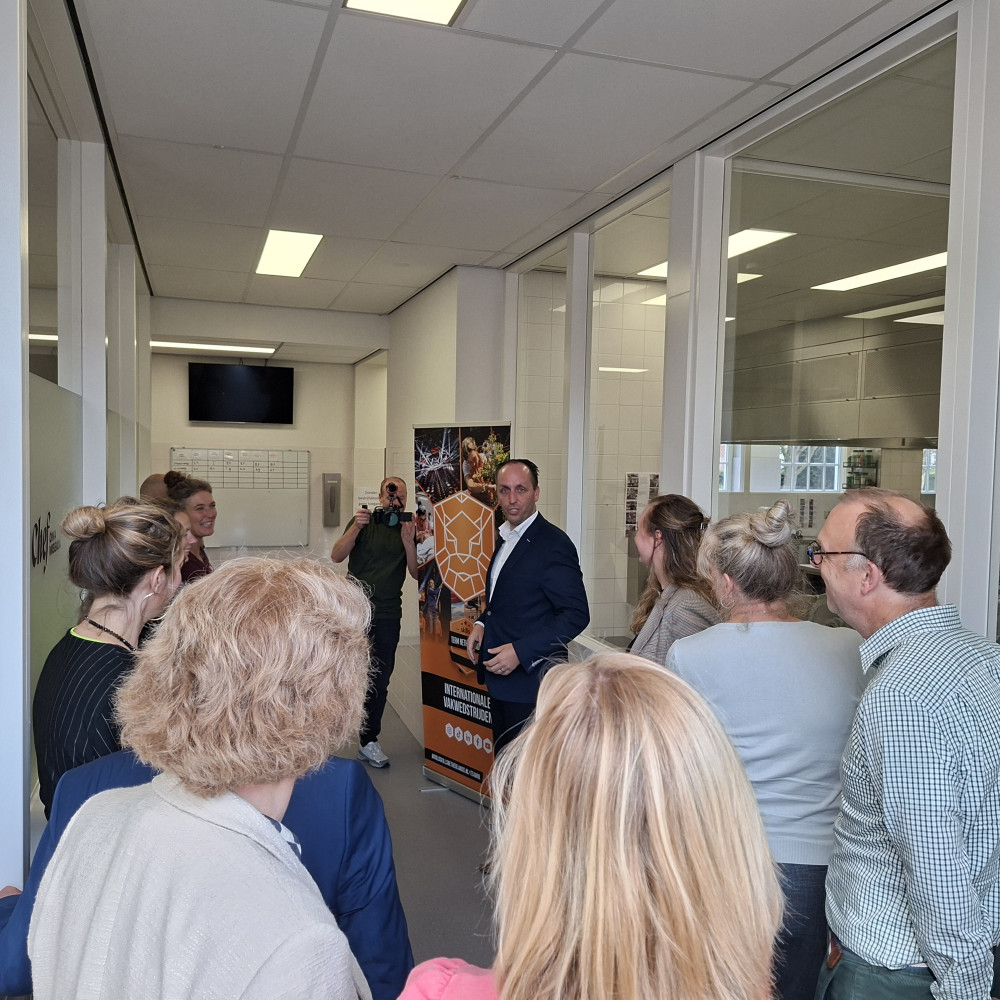 Groep mensen staat binnen rond een spreker die een rondleiding geeft door een gebouw van het Koning Willem I College.