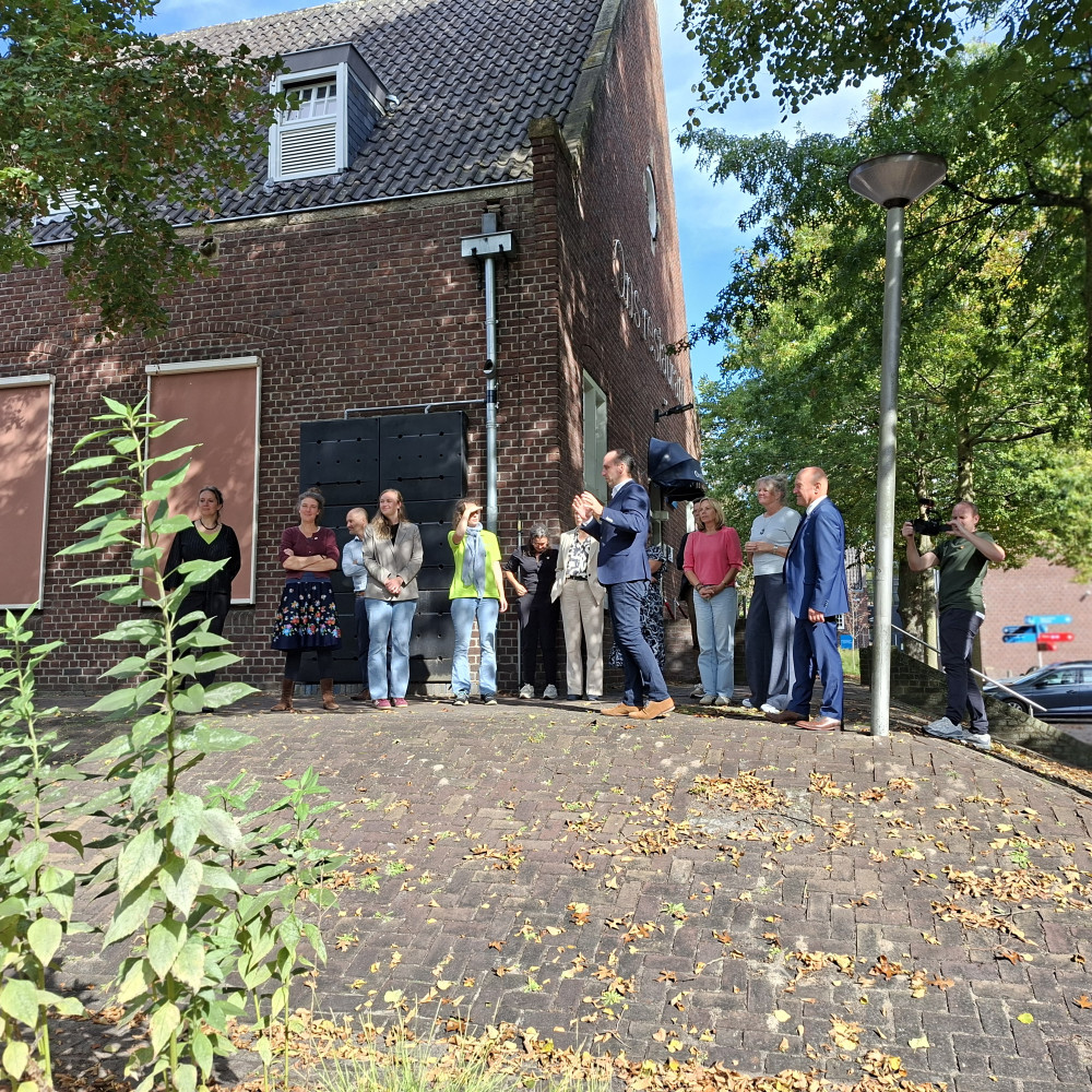 Groep mensen staat buiten bij een bakstenen gebouw op het terrein van Koning Willem I College en luistert naar een spreker, terwijl iemand de bijeenkomst fotografeert.