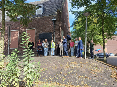 Groep mensen staat buiten bij een bakstenen gebouw op het terrein van Koning Willem I College en luistert naar een spreker, terwijl iemand de bijeenkomst fotografeert.