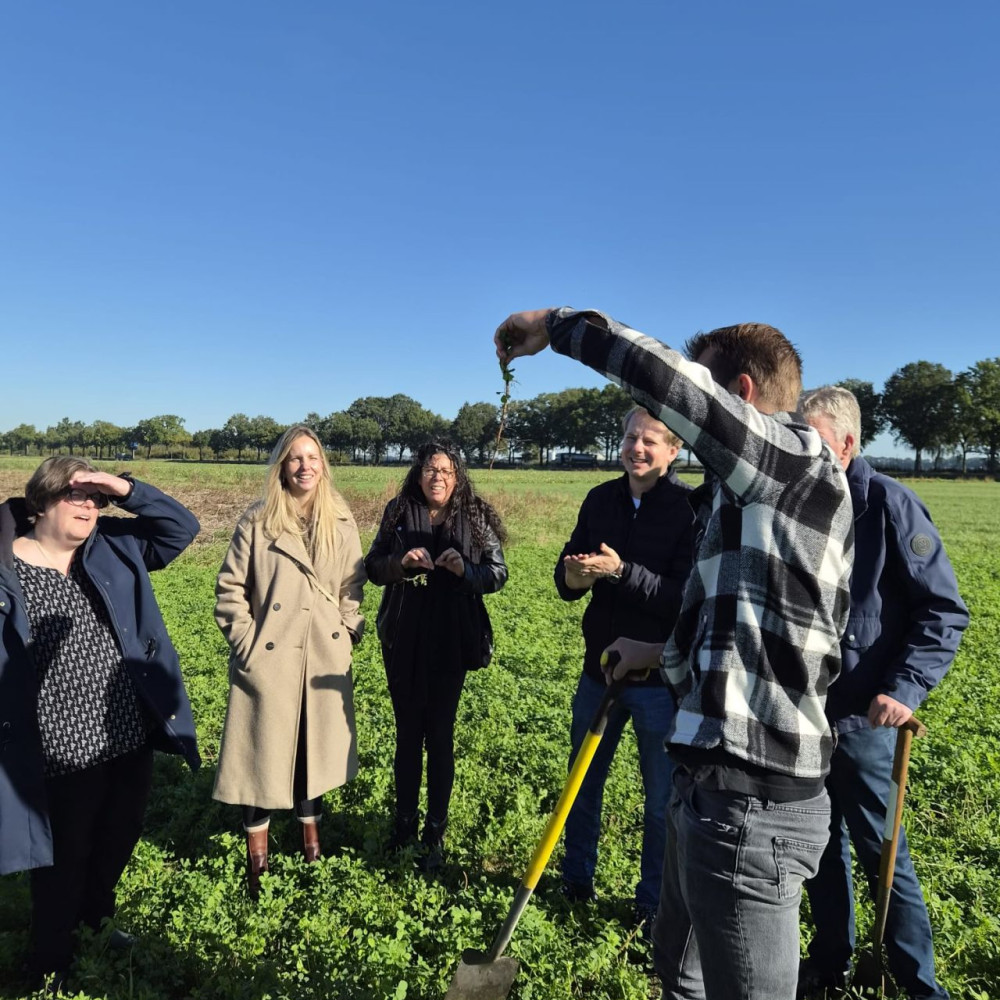 Een jonge boer houdt een plant met wortels omhoog op een akker met verschillende medewerkers van de provincie om hen heen.
