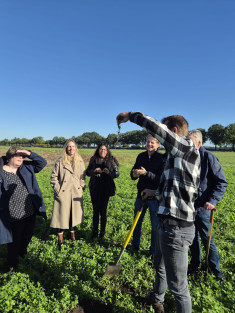 Een jonge boer houdt een plant met wortels omhoog op een akker met verschillende medewerkers van de provincie om hen heen.