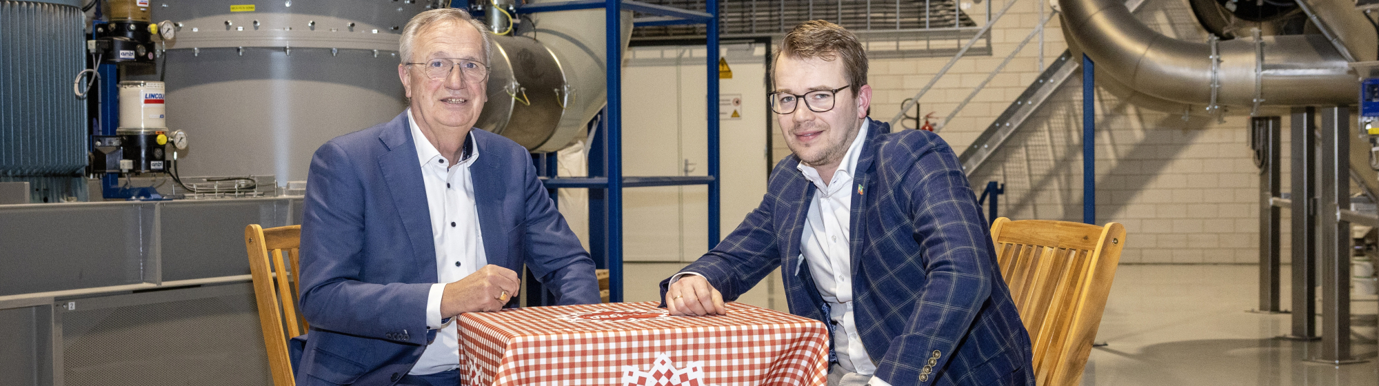 Jan Gijaarts en Henk Jan de Kat voeren overleg aan een tafel in een industriële omgeving met een rood-wit kleedje over samenwerking en innovatie in voedselverspilling.