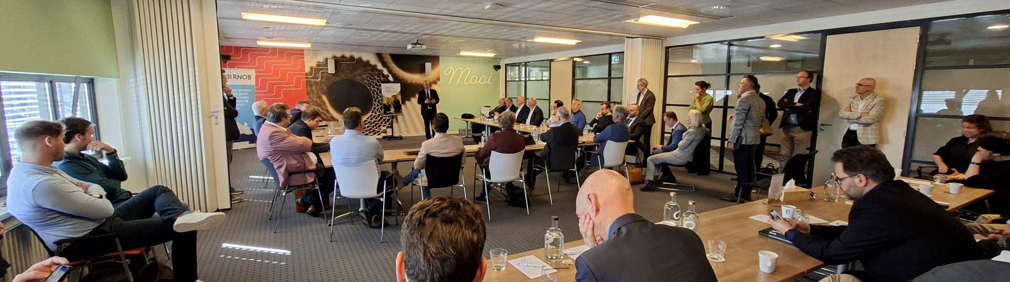 Wethouders en burgemeesters in een grote zaal, staand of zittend aan tafels, die luisteren naar een presentatie van een andere bestuurder..