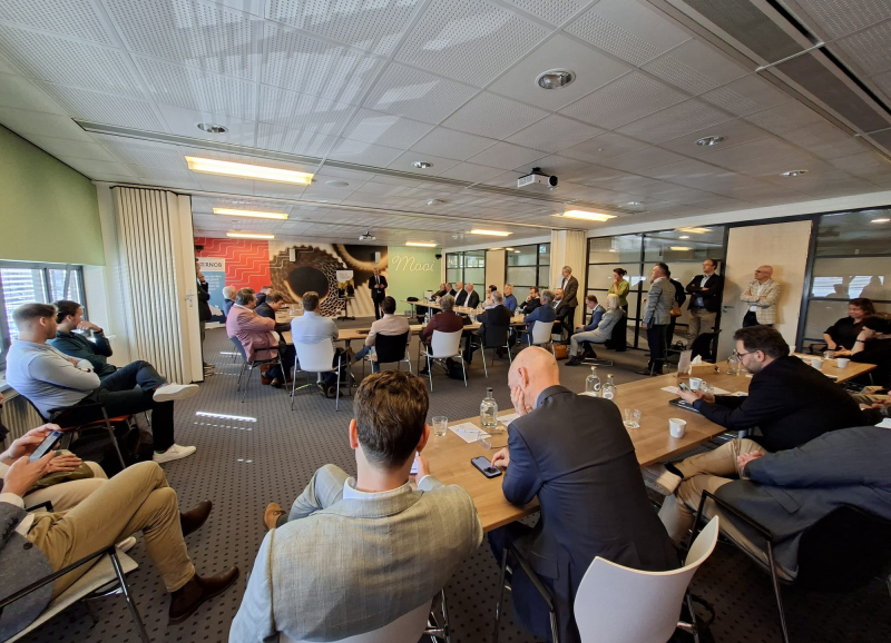 Wethouders en burgemeesters in een grote zaal, staand of zittend aan tafels, die luisteren naar een presentatie van een andere bestuurder..