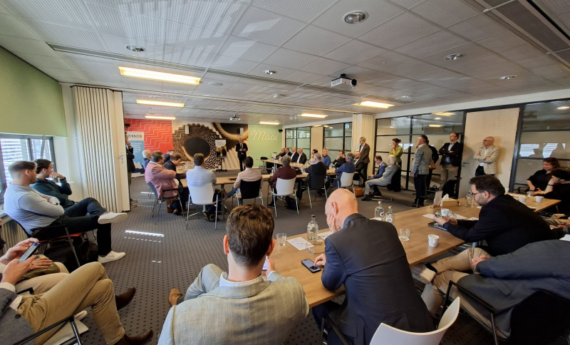 Wethouders en burgemeesters in een grote zaal, staand of zittend aan tafels, die luisteren naar een presentatie van een andere bestuurder..